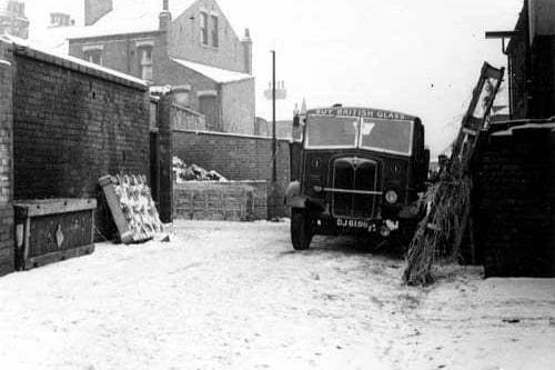 The yard of number 69, the Modern Art Glass Co. A lorry is parked next to some wooden glass crates with straw packing. The lorry has 'Buy British Glass' above its split windscreen. More crates are stacked on the left and rear of the yard, which is surrounded by a brick wall. Houses are visible behind. Snow is on the ground. Pictured in January 1945.