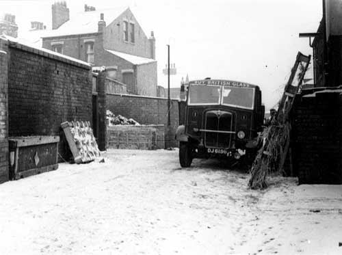 The yard of number 69, the Modern Art Glass Co. A lorry is parked next to some wooden glass crates with straw packing. The lorry has 'Buy British Glass' above its split windscreen. More crates are stacked on the left and rear of the yard, which is surrounded by a brick wall. Houses are visible behind. Snow is on the ground. Pictured in January 1945.
