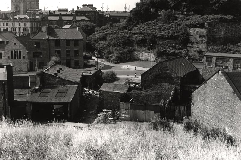 A photograph of Broad Chare taken in 1977. The view is looking down towards Broad Chare and Dog Bank. In the foreground there is a grassy bank which slopes down to the rear of buildings on Broad Chare and Cowgate. Trinity House is in the centre to the left with the entrance to Dog Bank then Cowgate to the right. Behind Trinity House are buildings on streets leading from the Quayside. Part of the Tyne Bridge can be seen in the distance. 