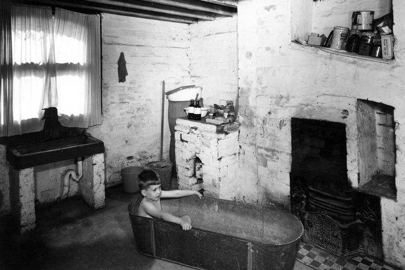 The scullery of a typical back-to-back house in Leeds, in this case being used as a bathroom. A sink can be seen on the left and a young boy is sitting in a tin bath in the centre. A fireplace is on the right but no fire appears to be lit.