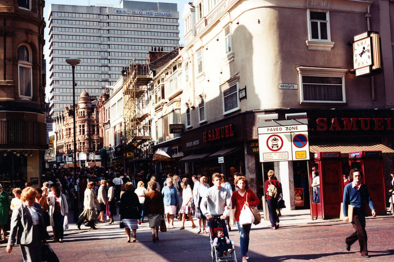 Looking along a crowded Commercial Street from Briggate. A sign indicates that this is a paved zone with no entry to vehicles. On the right, two red telephone boxes can be seen in front of H.Samuel, jeweller, which has a large clock on the wall outside. The shop just visible is Manfield footwear. Further up the street, scaffolding covers the front of a shop. West Riding House can be seen towering in the background.