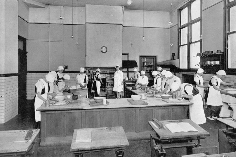 A cookery room in the girl's section of West Leeds High School circa 1940s. The pupils are dressed in white aprons and caps, and a range style cooker is visible at the back of the room.