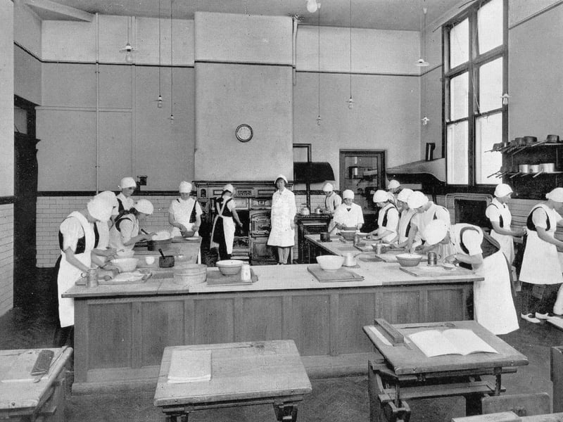 A cookery room in the girl's section of West Leeds High School circa 1940s. The pupils are dressed in white aprons and caps, and a range style cooker is visible at the back of the room.