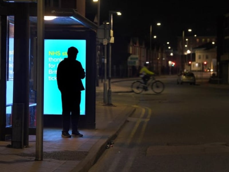 One lone traveller waiting at the bus stop, with a thank you NHS billboard behind him.
