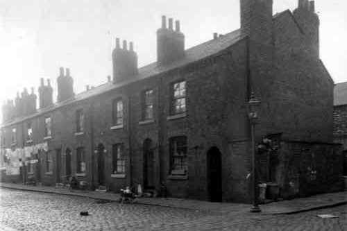 Odd numbered back-to-back properties on Bush Street, numbers run from the left in descending order to number 15 towards the right where a baby sits up in a pram. Next to this house on the right edge is a row of outside toilets and a storage area for dustbins. Pictured in September 1958.