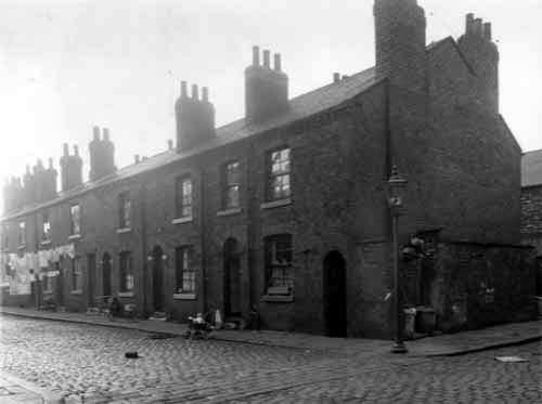 Odd numbered back-to-back properties on Bush Street, numbers run from the left in descending order to number 15 towards the right where a baby sits up in a pram. Next to this house on the right edge is a row of outside toilets and a storage area for dustbins. Pictured in September 1958.