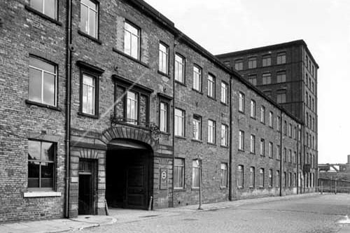 The arched entrance of Hunslet Mills at 23-25 Goodman Street. Most of the building is three storey with a seven storey section on the right. In the background is a railway line on top of an embankment. Pictured in September 1950.
