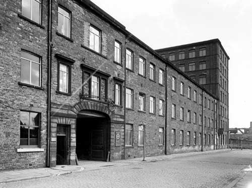 The arched entrance of Hunslet Mills at 23-25 Goodman Street. Most of the building is three storey with a seven storey section on the right. In the background is a railway line on top of an embankment. Pictured in September 1950.