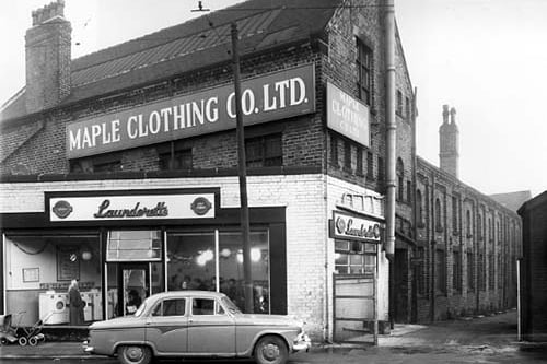 A car parked outside a Bendix, Self Service Laundrette in December 1958.. Lots of people and washing machines are visible through the windows and Christmas decorations are visible hanging from the ceiling. Two prams are visible waiting outside the laundrette on the far left. Above and behind the laundrette is the Maple Clothing Co Ltd, blouse factory. On the right is Admiral Street.