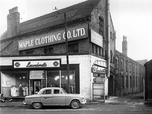 A car parked outside a Bendix, Self Service Laundrette in December 1958.. Lots of people and washing machines are visible through the windows and Christmas decorations are visible hanging from the ceiling. Two prams are visible waiting outside the laundrette on the far left. Above and behind the laundrette is the Maple Clothing Co Ltd, blouse factory. On the right is Admiral Street.