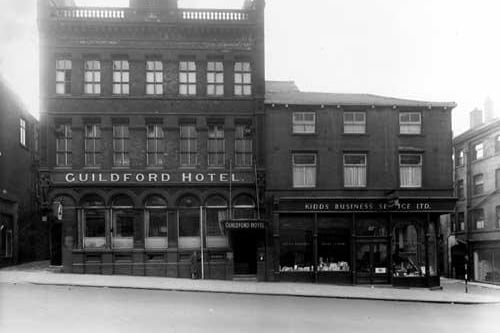 The south-west side of The Headrow showing number 115 The Guildford Hotel and number 117 Kidds Business Service Ltd in December 1949. A man stands in front with a measuring stick. To the far left is the entrance to Green Dragon Yard, to the far right is Basinghall Street. 