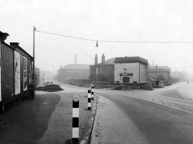 Crown Point Road looking south south west at the junction with Black Bull Street. Advertising hoardings on the left. Street light suspended on wire between two poles on either side of the street. Several striped bollards with reflectors on top positioned at the edge of the pavement. The premises of Edward Bidgood & Co, Black Bull Street can be seen in the distance on the left. Pictured in October 1953.