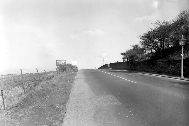 A view along Dewsbury Road facing north towards Leeds. Streetlamps on the right and a sign for Ediswan Mazda radio valves on the left. Field behind a barbed wire fence on the left. Pictured in October 1955.
