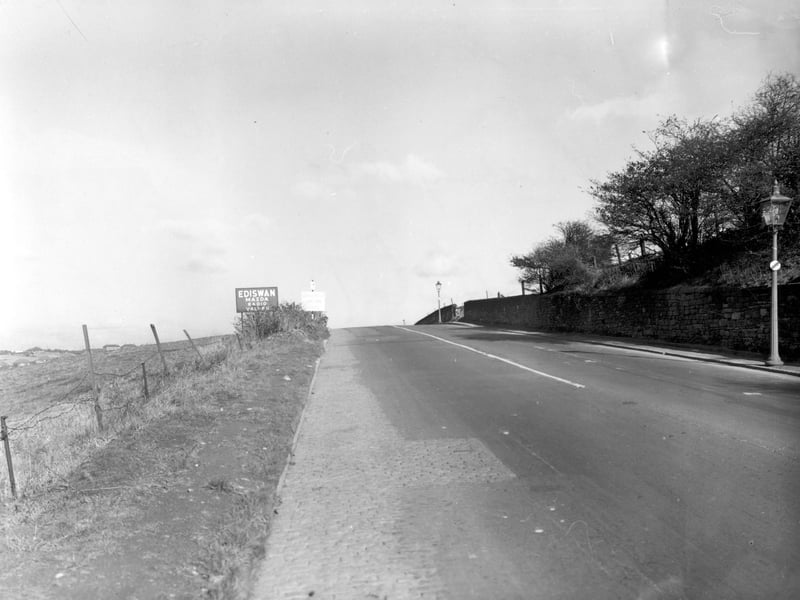 A view along Dewsbury Road facing north towards Leeds. Streetlamps on the right and a sign for Ediswan Mazda radio valves on the left. Field behind a barbed wire fence on the left. Pictured in October 1955.