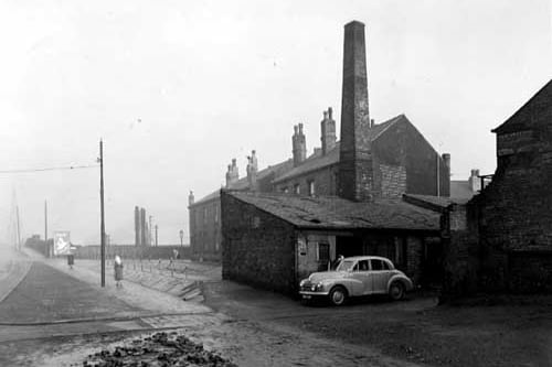 Looking north along Balm Road, with the entrance to Robinsons scrap metal yard. A Morris Oxford car is parked in the entrance with a man nearby. The low brick building has a large chimney. Houses are visible behind, as is a hoarding advertising Persil. Pictured in December 1951.