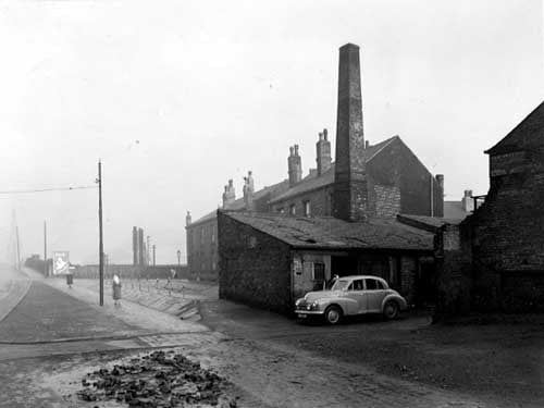 Looking north along Balm Road, with the entrance to Robinsons scrap metal yard. A Morris Oxford car is parked in the entrance with a man nearby. The low brick building has a large chimney. Houses are visible behind, as is a hoarding advertising Persil. Pictured in December 1951.