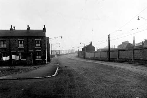 Looking south along Balm Road, with Back Barmouth Street on the left in Novembe 1955. Washing is hanging in some of these back yards. The road curves round to the right, is bordered by pavements and street lamps. On the right are tram lines; the lamp posts doubling as supports for the overhead power cables. A long wall is on the right with an isolated house at the end. In the middle of the photograph is a row of advertising hoardings. Open ground to the left is in front of Back Barmouth Street.