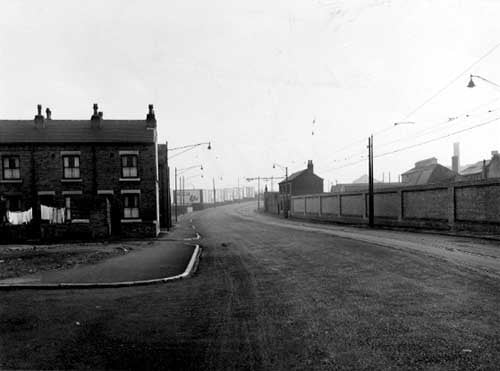 Looking south along Balm Road, with Back Barmouth Street on the left in Novembe 1955. Washing is hanging in some of these back yards. The road curves round to the right, is bordered by pavements and street lamps. On the right are tram lines; the lamp posts doubling as supports for the overhead power cables. A long wall is on the right with an isolated house at the end. In the middle of the photograph is a row of advertising hoardings. Open ground to the left is in front of Back Barmouth Street.