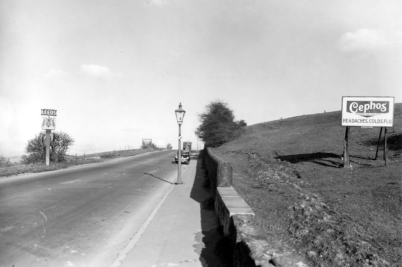 View along Dewsbury Road facing north towards Leeds. Leeds City coat of arms boundary sign and advertisements for Ediswan Mazda radio valves and Cephos cold and flu relief visible. A car and a truck drive towards the camera. A streetlamp can be seen. Pictured in October 1955.