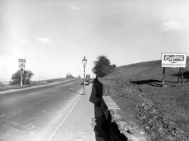 View along Dewsbury Road facing north towards Leeds. Leeds City coat of arms boundary sign and advertisements for Ediswan Mazda radio valves and Cephos cold and flu relief visible. A car and a truck drive towards the camera. A streetlamp can be seen. Pictured in October 1955.
