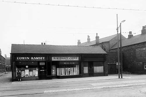 The odd numbered side of Wainwright Row is visible along the left edge of this view from April 1959. Then moving right is a row of 3 one storey shops. Number 53 on the left is Edwin Ramsey, electrical and radio engineer. In the centre, number 53a is a bakers and confectioners run by Harry Midgley and Sons. To the right number 55 is vacant, although directories list that it had been a dressmakers. Next right the garage or stable set back from the road is number 57 with the side of number 59, belonging to Ken Marsey, a plumber and contractor, visible on the right edge.