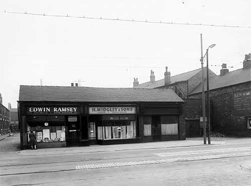 The odd numbered side of Wainwright Row is visible along the left edge of this view from April 1959. Then moving right is a row of 3 one storey shops. Number 53 on the left is Edwin Ramsey, electrical and radio engineer. In the centre, number 53a is a bakers and confectioners run by Harry Midgley and Sons. To the right number 55 is vacant, although directories list that it had been a dressmakers. Next right the garage or stable set back from the road is number 57 with the side of number 59, belonging to Ken Marsey, a plumber and contractor, visible on the right edge.