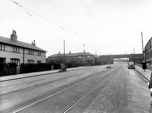  Belle Isle Road looking south towards an iron railway bridge in November 1950. On the right of the road there is a brick industrial building with a wall and iron fence running along the pavement. There is a woman walking along the pavement and a car parked at the kerb. On the left are two rows of stone terraced houses. In the road are tram lines and a traffic island.At the left hand kerb stands a window cleaners barrow.
