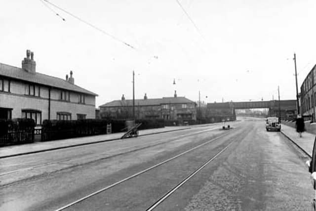 Belle Isle Road looking south towards an iron railway bridge in November 1950. On the right of the road there is a brick industrial building with a wall and iron fence running along the pavement. There is a woman walking along the pavement and a car parked at the kerb. On the left are two rows of stone terraced houses. In the road are tram lines and a traffic island.At the left hand kerb stands a window cleaners barrow.