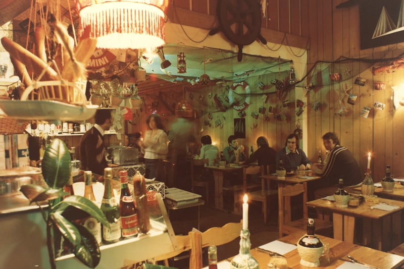 A photograph of the interior of Marco Polo Italian restaurant taken in 1976. The restaurant is decorated in a nautical style with fishing nets shells lobsters etc. The tables and chairs are made of pine additional lighting is provided by candles in empty wine bottles. The walls are decorated with pine cladding. 