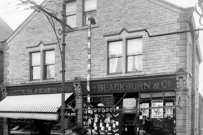 Town Street in May 1906. On the left is number 182, business of John Myers, greengrocer, next number 180, Willaim Blackburn and Co, clothiers. The window has a display of goods, in the right window are posters for 'Sailor Suits 2/6d' (12 1/2p), 'Belted Suits' 4/11d (just under 25p). Workmen are measuring the building, boys in front watching the photographer.