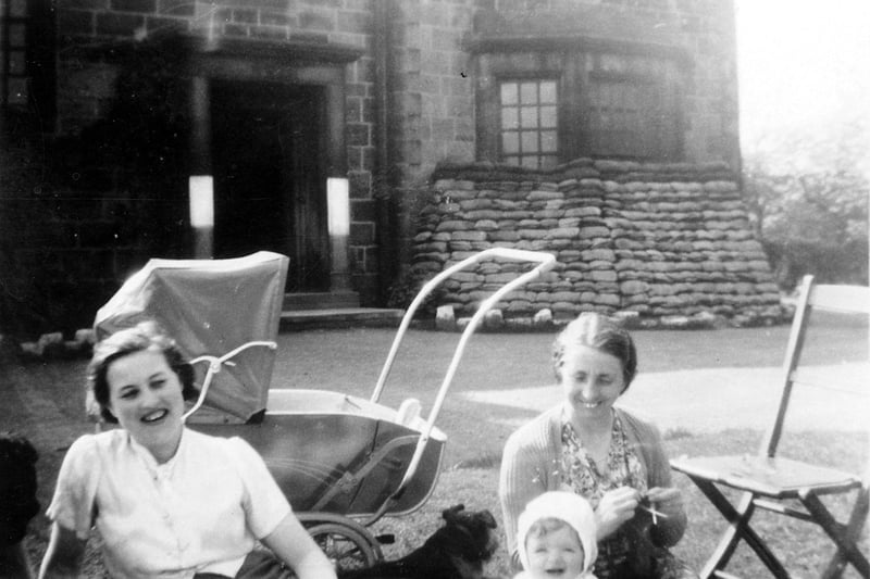 Photograph of Morley Hall Maternity Home, taken during the Second World War when sandbags were piled up in front of the building to protect it from bomb damage. Two young women are sitting in the garden with a baby, a dog and a pram.