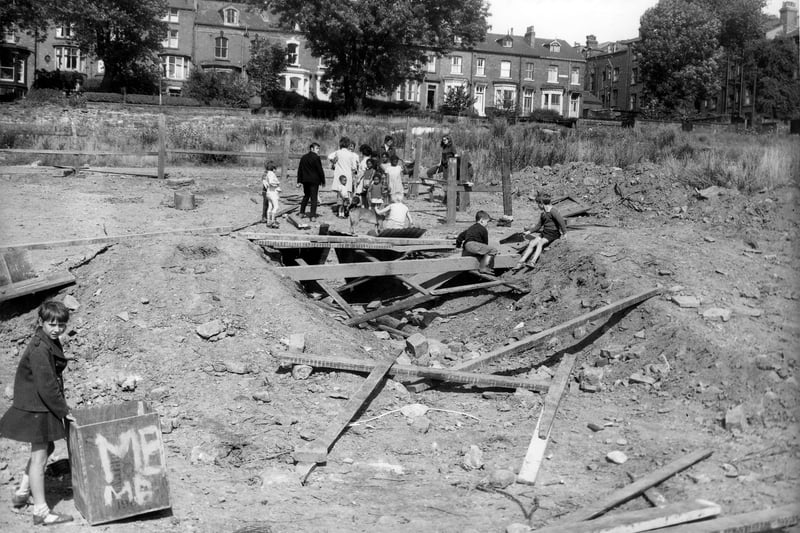 Running from left to right in the background of this view are houses on St. John's Grove in August 1969. In the foreground is an area of wasteland, now the site of houses which had stretched from Hyde Park Road to Woodsley Road. The land is being used as an adventure playground. Several children sit on a makeshift bridge made of wooden planks. A girl holds a tea chest in the bottom left.