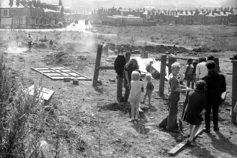 An area of wasteland being used as a children's playground. In the background to the left are houses on Woodsley Road with Hyde Park Road on the right. In the centre is the Sacred Heart Church with its concrete structure and flat roof. In the foreground children, children use wood and corrugated iron to make playground apparatus supervised by several adults. Pictured in August 1969.