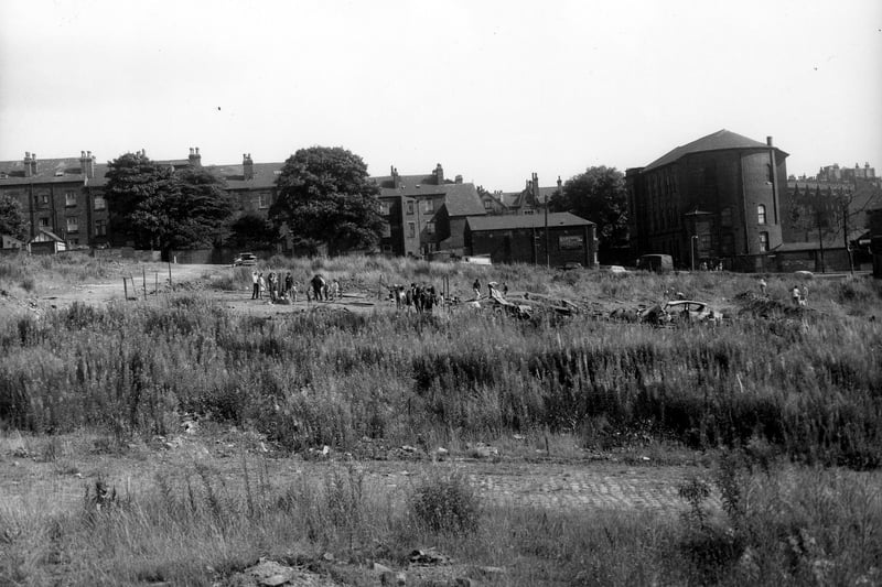 The site of back-to-back streets demolished as part of Leeds slum clearance programme. In the foreground a line of stone setts can just be made out, this was the site of Osbourne Street. At the time of this view, the land was being used as an adventure playground. In the centre a large group of children and adults construct temporary play apparatus from planks of wood and sheets of corrugated iron. Houses on St. John's Avenue can be seen in the background to the left with premises of Harp Mill Paint and Varnish Co. Ltd. in the centre at No. 65 Woodsley Road. On the right is the Belle Vue Methodist Church.