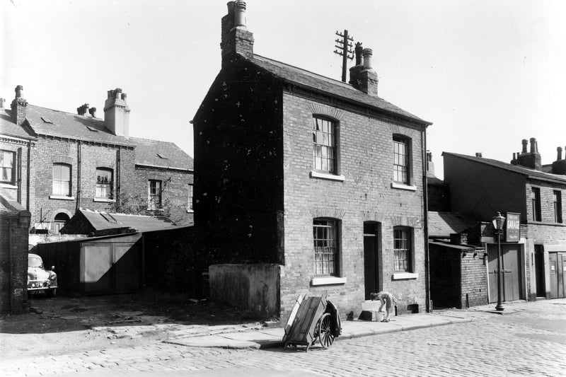 This view looks across properties of West End Terrace in August 1961. In the background on the left are properties on Cliff Road. Number 23 West End Terrace is visible in the centre with a wooden handcart outside. A lady cleans the front steps. On the right edge is the West End Garage at number 24.