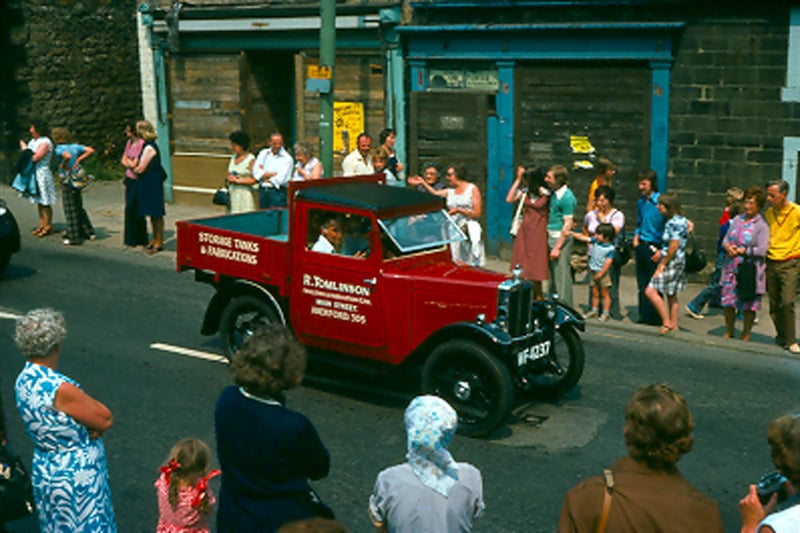A colour photo of a vehicle belonging to R. Tomlinson forming part of a parade of floats travelling along Upper Town Street during the Bramley Carnival of 1976. People line the streets to watch the newly resurrected carnival, the first since the early part of the 20th century. Shops in the background at no. 220 and 222 are boarded up and soon to be demolished.