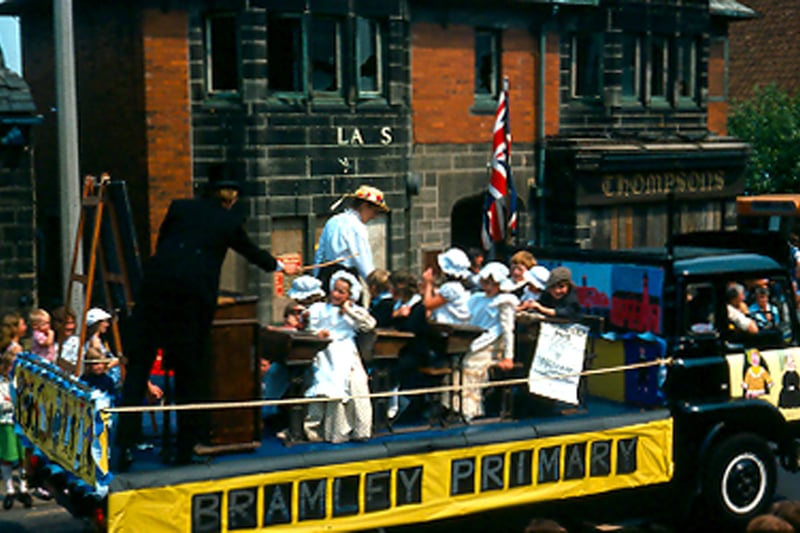 Bramley Primary School's float in the Bramley Carnival of 1976. Teachers and pupils are dressed in Victorian costume and are depicting a Victorian school day. The float is part of a parade travelling along Upper Town Street; shops in the background, including Thompson's bakers and confectioners at no. 216, are boarded up and scheduled for demolition.