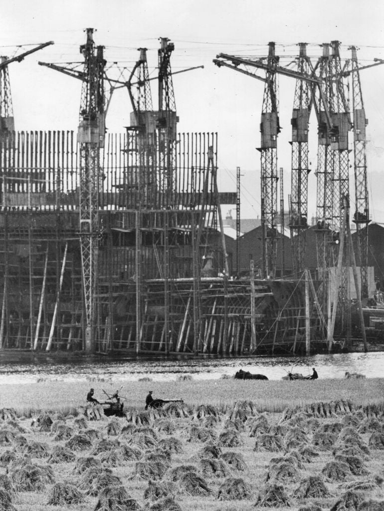Work proceeds on the sister ship for the 'Queen Mary'; 'Cunarder 552', as she is known, may be seen rising up between the scaffolding on the River Clyde.  