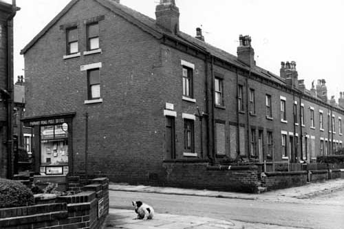View looks from the end of Back Westbury Street onto Parnaby Road in July 1967. On the left is the Parnaby Road Post Office at number 29 Parnaby Road. In the foreground, a black and white dog scratches its ear. Properties on the even numbered side of Parnaby Mount are visible to the right.