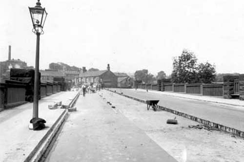 Resurfacing of Kirkstall bridge in June 1938. Men working on the road, wheelbarrow & broom on road. Lamppost on left hand side with some clothing piled up at its base. Kirkstall Brewery & tall chimney in background.