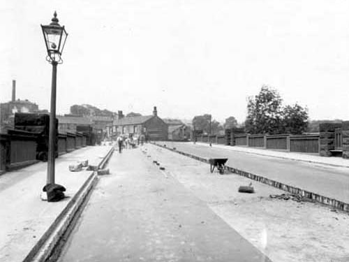 Resurfacing of Kirkstall bridge in June 1938. Men working on the road, wheelbarrow & broom on road. Lamppost on left hand side with some clothing piled up at its base. Kirkstall Brewery & tall chimney in background.