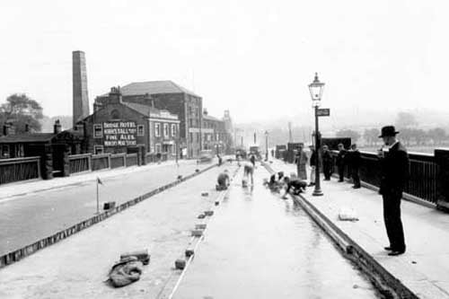 Re-waterproofing decking on Kirkstall Bridge. men are working on road which is partially dug up and under water. Several men are watching. Bridge Hotel & tall chimney on left. Lamp post also LCT bus stop on right. Pictured in June 1938.