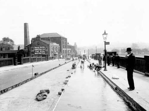 Re-waterproofing decking on Kirkstall Bridge. men are working on road which is partially dug up and under water. Several men are watching. Bridge Hotel & tall chimney on left. Lamp post also LCT bus stop on right. Pictured in June 1938.