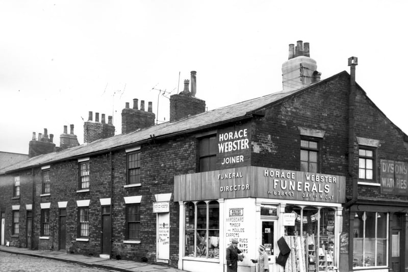 Whitby Street with two businesses on Armley Road in the foreground. In focus are the premises of Horace Webster Funeral Directors, also advertising lino, carpets, rugs etc & hardboard and timber. Rolls of lino are displayed outside the shop front where a couple stand talking.