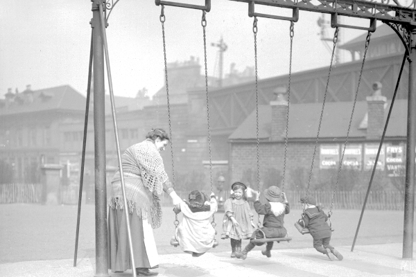 Down the Gorbals shortly after the turn of the 20th century, a woman swings her children from a Victorian-era swingset
