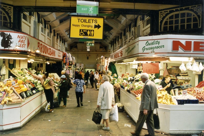 Shoppers and stalls inside Kirkgate Market Hall in September 1999. On the left is Brooksbank Greengrocers and on the right is Neil's Greengrocers. A sign is positioned to the centre of the photograph showing the direction of the Toilets and Nappy Changing area.