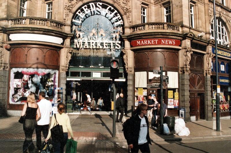 The entrance to the indoor market on Vicar Lane in September 1999. A Pelican crossing, pedestrians and street signs can be seen. A newsagent and computer game shop are to the right. A newspaper seller is partially visible.