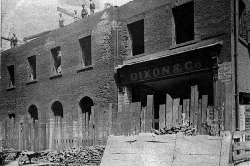 Bishopgate Street improvements. A partially demolished building is main view. At the top corner of the block is a shop with the name Dixon and Co. over the top. Poster and playbills are stuck on the walls. Workmen are standing on the roof of the building. Pictured in June 1899.