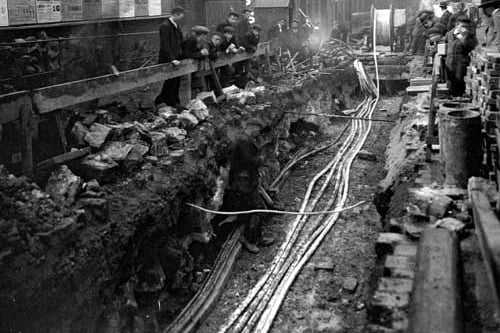 A view along the length of excavations of old cellars in Aire Street, showing piping being laid, workman standing at edge of trench and workman's hut. Also in view is a wall covered in various bill posters, gas lamp and premises of Rhodes and North. Pictured in May 1899.