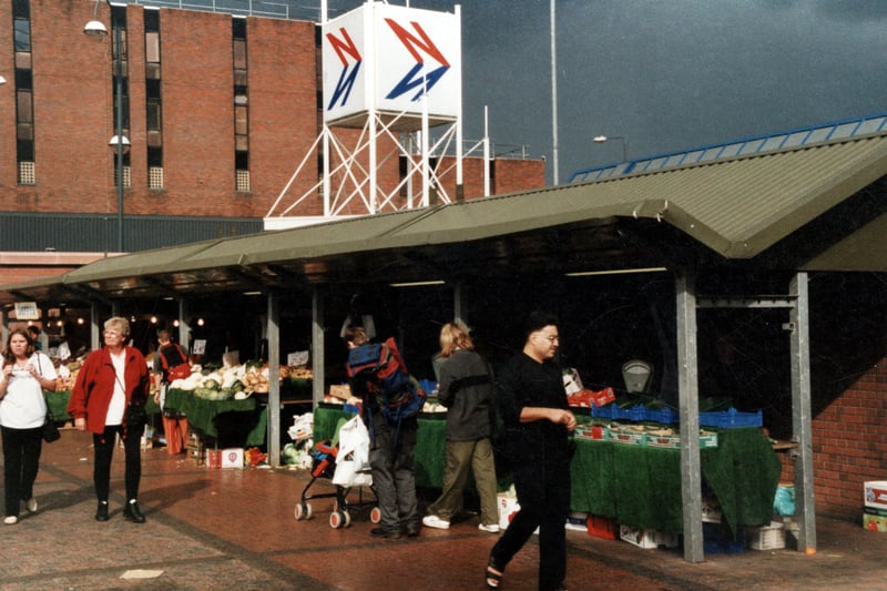 This view of the outdoor market shows a row of fruit and vegetable stalls. In the background is the Leeds City bus station with the National Express sign prominent. In the distance is Millgarth Police Station. Pictured in September 1999.